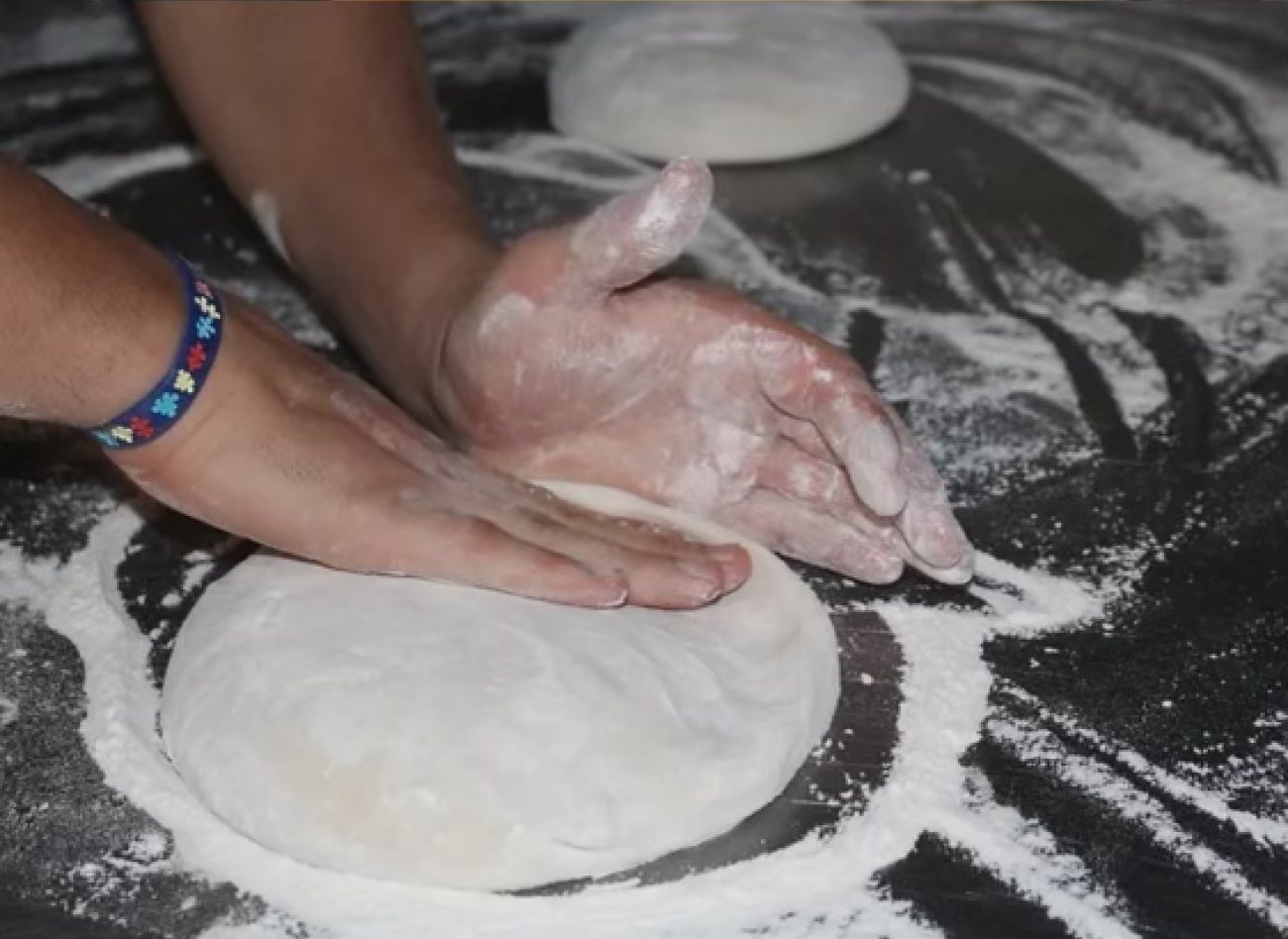 Two floured hands pressing a ball of pizza dough flat on a dark, flour-dusted bench.