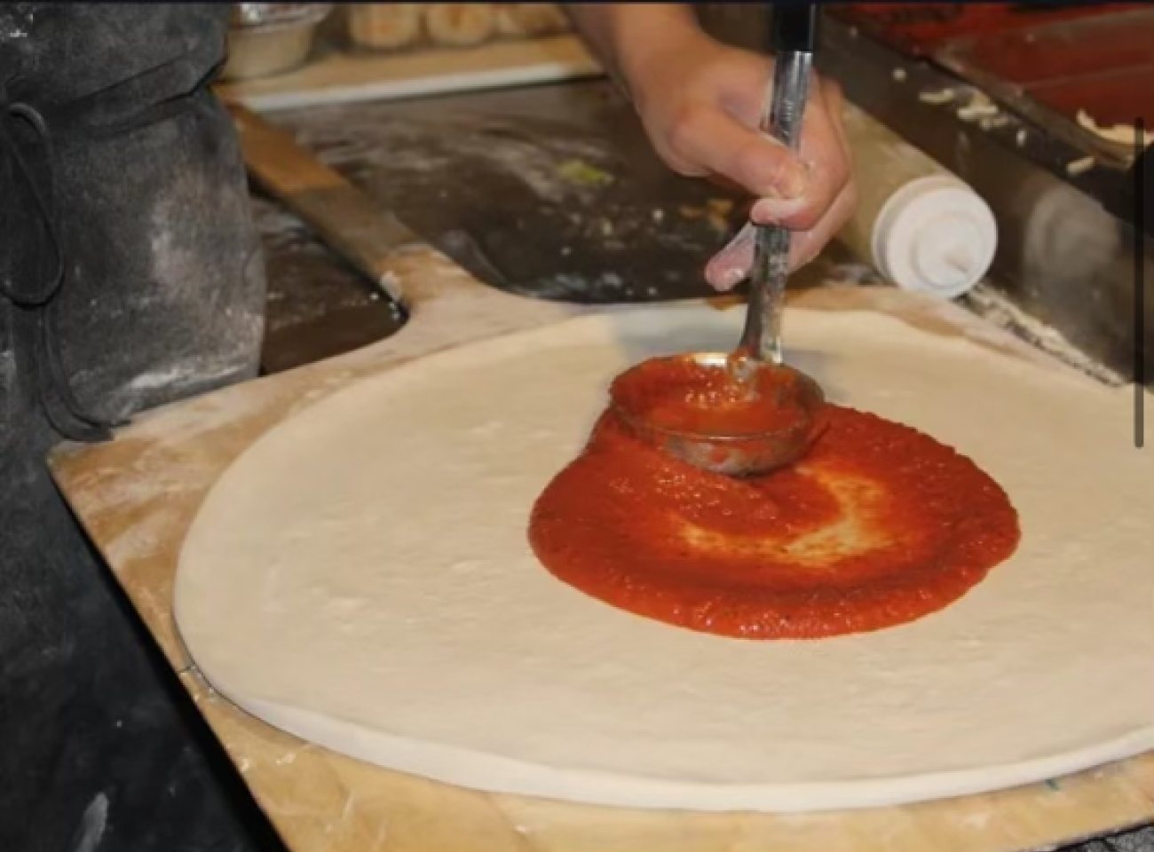 A ladle spreading red sauce in a circle across a raw, stretched pizza base on a wooden peel.